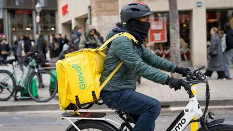 A migrant seen working as a rider for a delivery company in Barcelona. His face is half covered by scard, and he is wearing a helmet, a green puffer jacket and a yellow food delivery boxbag that says 'Glovo'.