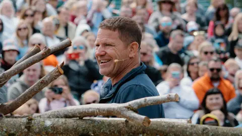 Steve Backshall CBBC presenter Steve Backshall with short brown hair who is wearing a blue top laughs as he gives a talk to a large crowd of people. He is standing in front of branches.