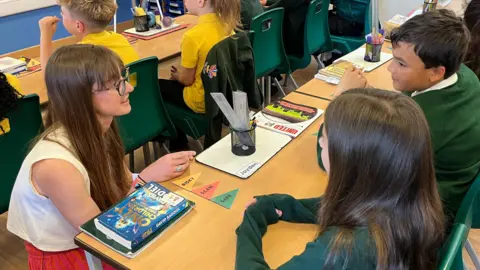 Miss Mancini is kneeling in front of a desk in a classroom talking to two children.