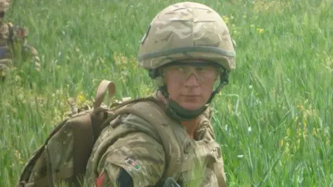 Luke Shipman Luke is in combat gear in a field. He is wearing full camouflage suit, including bag and hat