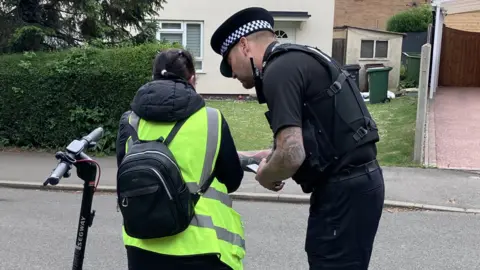 A police officer stands next to a woman seen from behind, as they discuss her e-scooter. She is wearing a high-vis neon vest and grey jogging trousers.