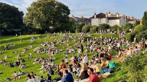 BBC/Steve Mellen A green park with many people, with a tree and houses in the background.