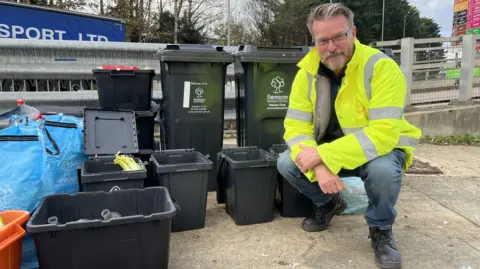 Councillor Chris Watts in a Swindon Borough Council high visibility jacket crouching next to recycling bins and bags.