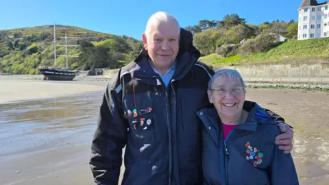 John and Mary Whelan stand together on the sandy beach. John, on the left, has his arm around Mary. They're both wearing dark blue coats, each with a collection of poppy badges on. They're an older couple and are smiling, standing in front of the grounded tall ship in the background on the left, with a hilly tree-lined coastline behind it.