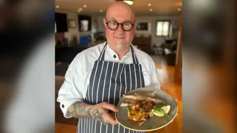 Nick Jaye is a bald, white man with circular black glasses on and a white soul patch. He is wearing a white chef's shirt under a stripy navy and white apron. He has tattoos covering his right arm and he is holding a grey plate with bright coloured vegetables and fish on it.