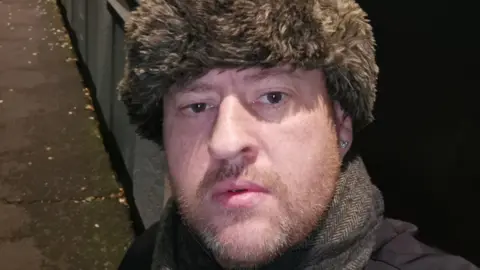 Spindrift A man, Gary Mooney, wears a fur lined russian hat and looks intensely into the camera 