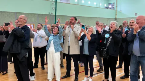  A crowd cheers and claps in a sports hall, with many wearing sky-blue rosettes.