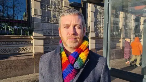 Mal O'Hara, who has greying short hair and a ginger/grey beard stares ahead to the camera while wearing a grey coat and rainbow coloured scarf. He is standing beside a bus shelter