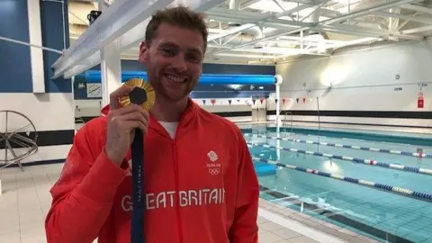 A man with blonde hair wearing a red jacket saying 'Great Britain' on it, holding up a gold medal. He's in a leisure centre and there's a swimming pool in the background.