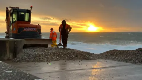 Two people on a beach with heavy machinery clearing the shingles from the promenade. The waves on the right are crashing onto the beach. 