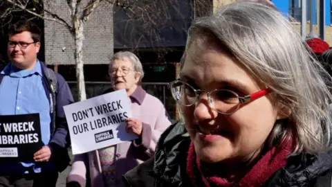 John Fairhall/BBC Rachel Wood is standing in front of protesters. she is wearing a read scarf and glasses. In the background you can see placards being held saying "don't wreck our libraries".  