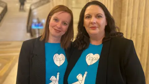 Two women both wearing black blazers with blue tops that show have a graphic of the outline of a child holding a balloon, in the balloon it says hope. The woman on the left has brown hair and the one on the right has black. 
