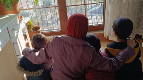 Nilobar wears a red hijab and a purple jacket and puts her arms around her three children, all wearing winter jackets - their backs to the camera as they look outside, sitting on the floor and looking out the window.   