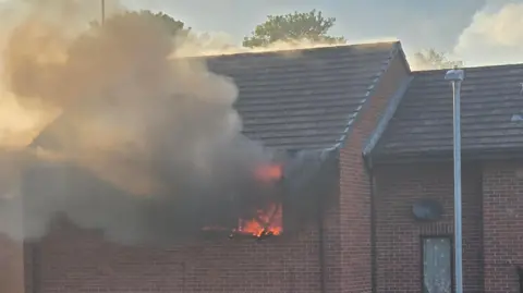 The upper floor of a brick residential building. A fire is blazing inside a window and thick smoke is billowing out.