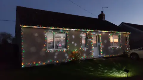 Luke McGann A bungalow is pictured at night with multi-coloured Christmas lights around it. Other lights are projected onto the garden grass in front of the home. 