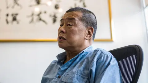 Jimmy Lai wears a blue denim shirt and sits on an office chair in front of a framed piece of Chinese calligraphy
