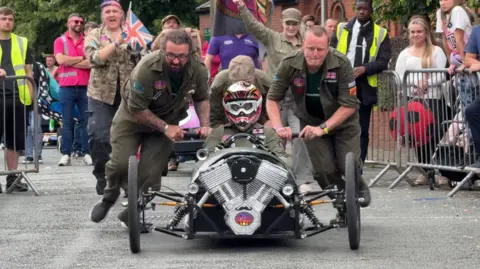 A team of four people push and drive a kart made to look like a racing car. The team and two women cheering them on are wearing military fatigues. Further supporters and stewards watch on from behind metal barriers. 