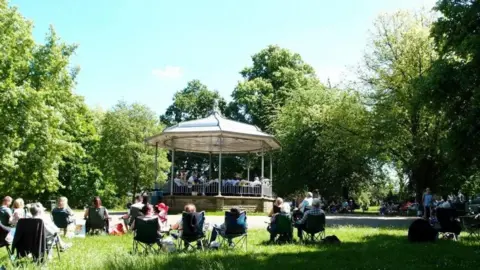 Ilkeston Brass A grassy park with many trees and a bandstand in the middle. The bandstand has a brass band on it. People are sat around the bandstand on fold out chairs.