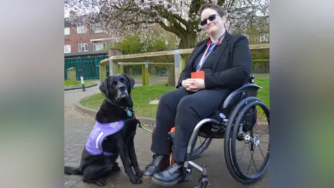 A woman sits in a wheelchair and smiles. She holds the lead of a black Labrador, which wears a purple vest.