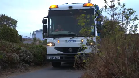 A bin lorry is seen driving down a country road with amber lights flashing. 