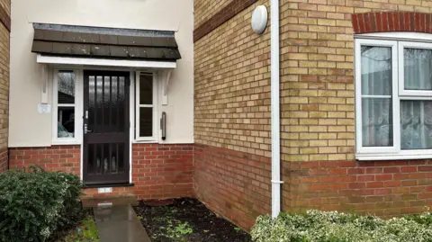 John Devine/BBC The outside of a ground floor flat, with hedges and shrubs growing at the front of it. There is a brown communal front door and the Chapmans live in a one-bedroom ground-floor flat on the right.