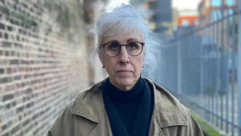 Nicola looks down the barrel of the camera with an alleyway in the background, looking sullen with her mouth closed. She has white hair, wears glasses and has a blue scarf underneath her khaki-coloured overcoat