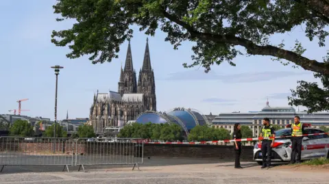 Three employees from the public order office stand on a street in Cologne next to a police cordon, with a Cologne cathedral in the background 