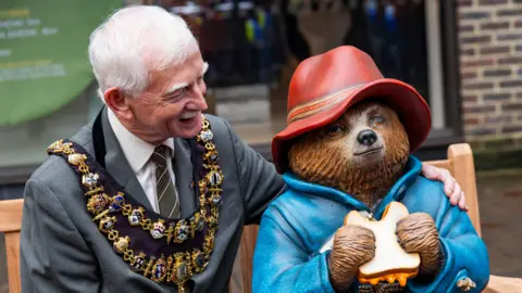 StudioCanal Mayor of Newbury Andy Moore - wearing a grey suit and with his ceremonial gold chain round his neck - has his arm around the statue of Paddington Bear at its unveiling ceremony 