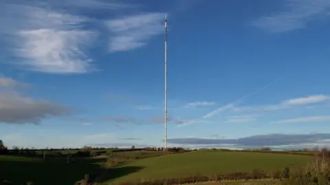 A single, towering mast rises straight from the rolling green fields, stretching high into a clear blue sky. Thin support wires angle out from its sides, emphasising its immense height. Small buildings at the base appear tiny in comparison. The mast stands sharply against the landscape, a tall, slender landmark dominating the horizon.