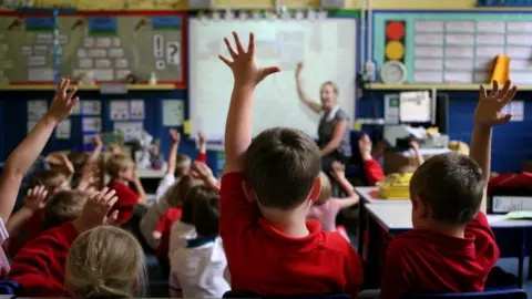 PA Media Young children in a classroom wearing red and white tops. Some have their hands up while a teacher is at the front of the room in front of a white board.

