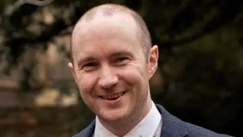 Robert Rees-Hughes smiling at the camera with short dark hair, stubble and wearing a suit outside. 