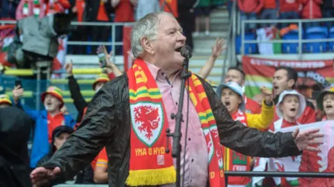 Dafydd Iwan, soaked in the rain and wearing a Wales scarf and a pink shirt, stands in a stadium singing into a microphone surrounded by Wales football fans singing Yma o Hyd
