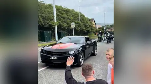 A black supercar leading the special vehicle parade in Bury followed by a number of motorbikes on a main road. It is a dry day and hills and the countryside can be seen in the background.