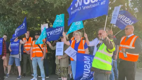 A group of teachers stand in a picket line outside Westbourne Academy. They hold banners and flags from their unions and some wear bibs and hi-vis jackets. 