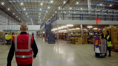 DP World A man in a pink hi-vis jacket walks through a large open depot. Around him are staff in hi-vis and white robots, as well as thousands of boxes.