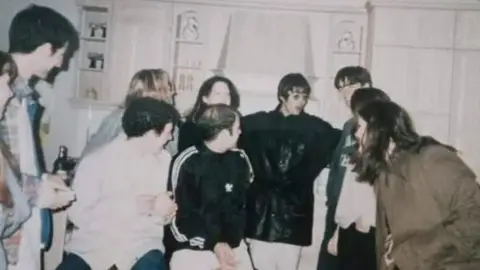 Abbie Eales Liam Gallagher wearing glasses and a large black jacket, surrounded by young people in a kitchen, with Bonehead. Everyone is looking at Liam