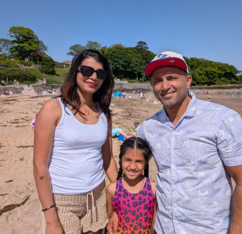 Three people - a man, woman and younger girl - stand close together on a sandy beach on a sunny day, posing for a photo. Behind them, other visitors sit and walk along the shoreline, with a low stone wall, grassy hillside, and houses partially visible. The sky is bright and clear.