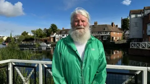 A man with a long grey beard wearing a green raincoat is standing in front of a river, there are boats docked behind him and some buildings visible in the background