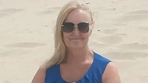 A woman with long blonde hair, sunglasses and a blue sleeveless top, smiles as she stands on a beach.