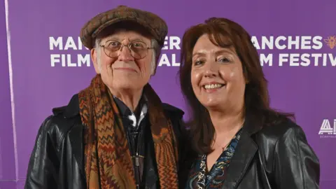 Suzan Holder wearing black on the right is smiling at the camera. Noddy Holder, who has a cap and scarf on, is on the left and they are both in front of a mainly purple background.