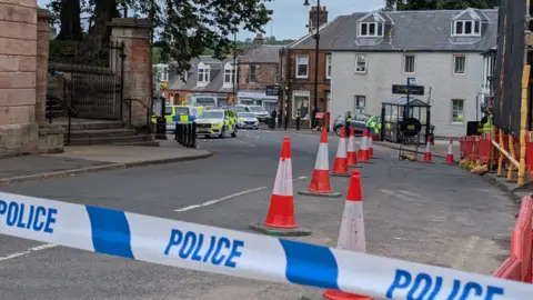 police tape seals off a road which has several police cares parked outside a row of houses