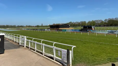 A wide image of the racecourse, showing a long white rail in the foreground on the left and other rails in the background. Trees are in the distance and blue sky features in the top third of the photo.