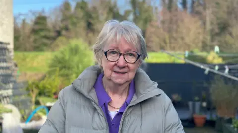 Kathline Kelly. She has short grey hair and a fringe, wearing glasses and a grey puffer coat with purple polo top underneath. A garden area is blurred behind her.