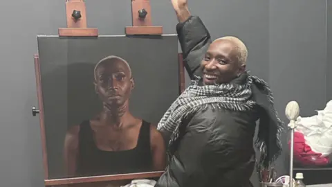 Ada Osigwe Ada Osigwe grins as she poses next to the portrait of herself. She has short cropped hair and wears a checked black and white scarf and dark coat. Her portrait shows her wearing a strapped black top and is seen with the keloid scar on her neck and chest.