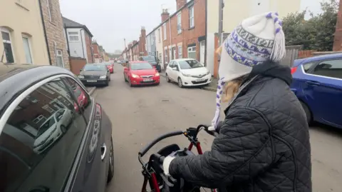 A woman wearing a white and purple patterned hat and black quilted coat is pushing a walking frame as she crosses the road to reach the path. A moving red car with headlights on faces her, leaving her in the centre of four stationary cars and one in transit trying to get past her.