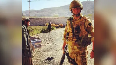 Doug Beattie A man is stood in full camouflage army fatigues, with helmet, multiple pouches and bags attached to him. He has a rifle in his right hand. He looks directly at the camera, to his right is a man wearing traditional afghan men's clothing. Behind him is a dirt road, a white wall and a large mountainside. 