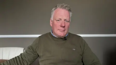 Man with grey hair looking into camera, wearing green/brown round neck jump