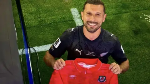 Kosta looking up from the pitch in his black New Zealand football kit and holding a red Umbro Wales jersey