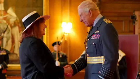 PA Media A woman with red-coloured hair wearing a black and white hat and black blazer shakes King Charles' hand. He is wearing a dark-coloured jacket.