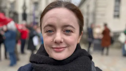 Una Boyd outside court. She is wearing a black scarf and black jacket. Her brown hair is tied back. There are people behind her.